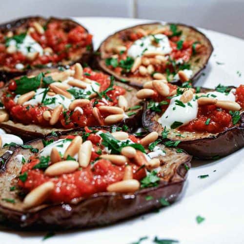 A platter of sliced and roasted eggplant topped with tomato sauce, yoghurt and roasted pine nuts with a final garnish of chopped parsley.