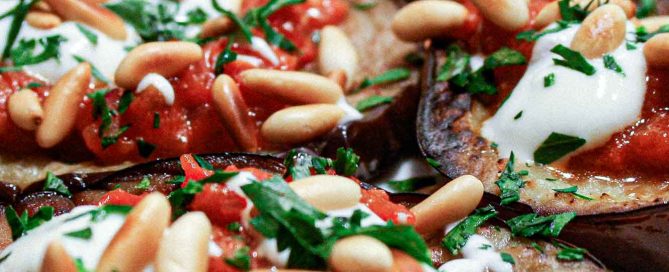 A platter of sliced and roasted eggplant topped with tomato sauce, yoghurt and roasted pine nuts with a final garnish of chopped parsley.