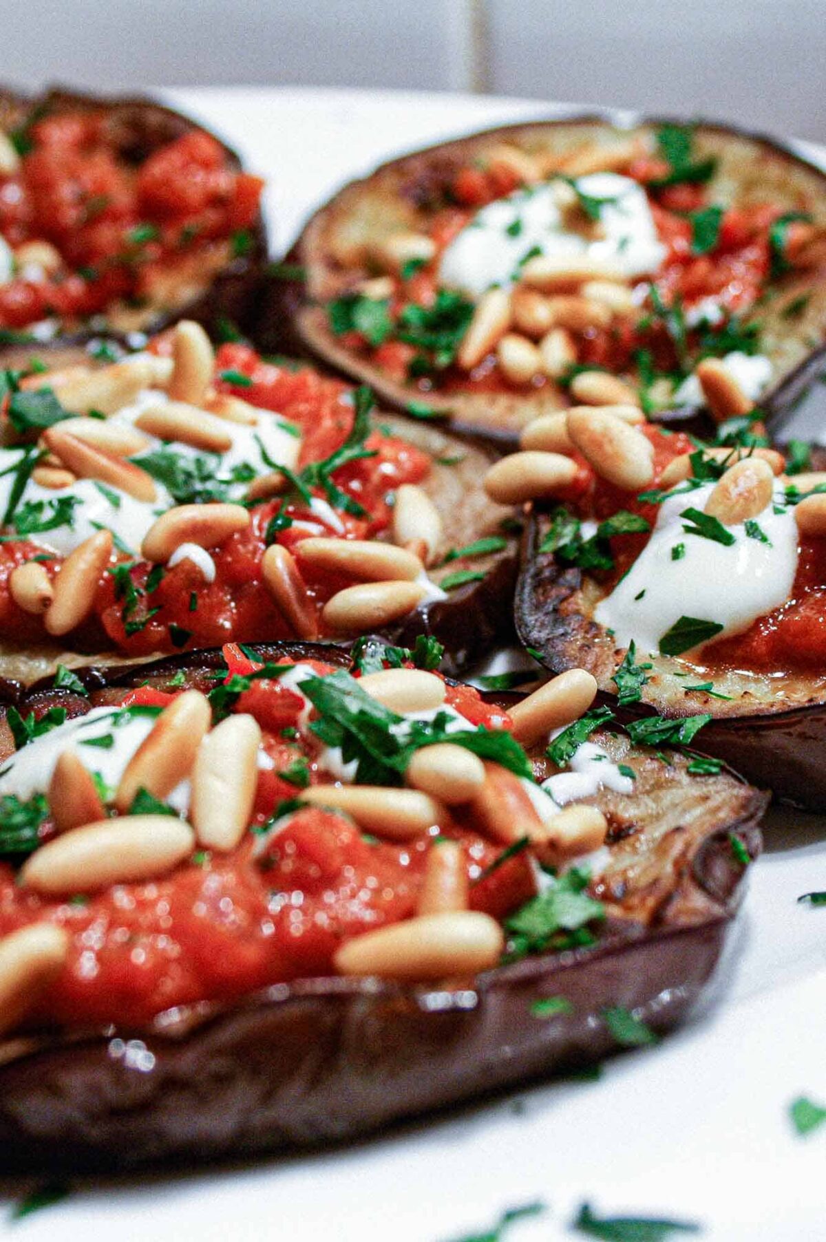 A platter of sliced and roasted eggplant topped with tomato sauce, yoghurt and roasted pine nuts with a final garnish of chopped parsley.