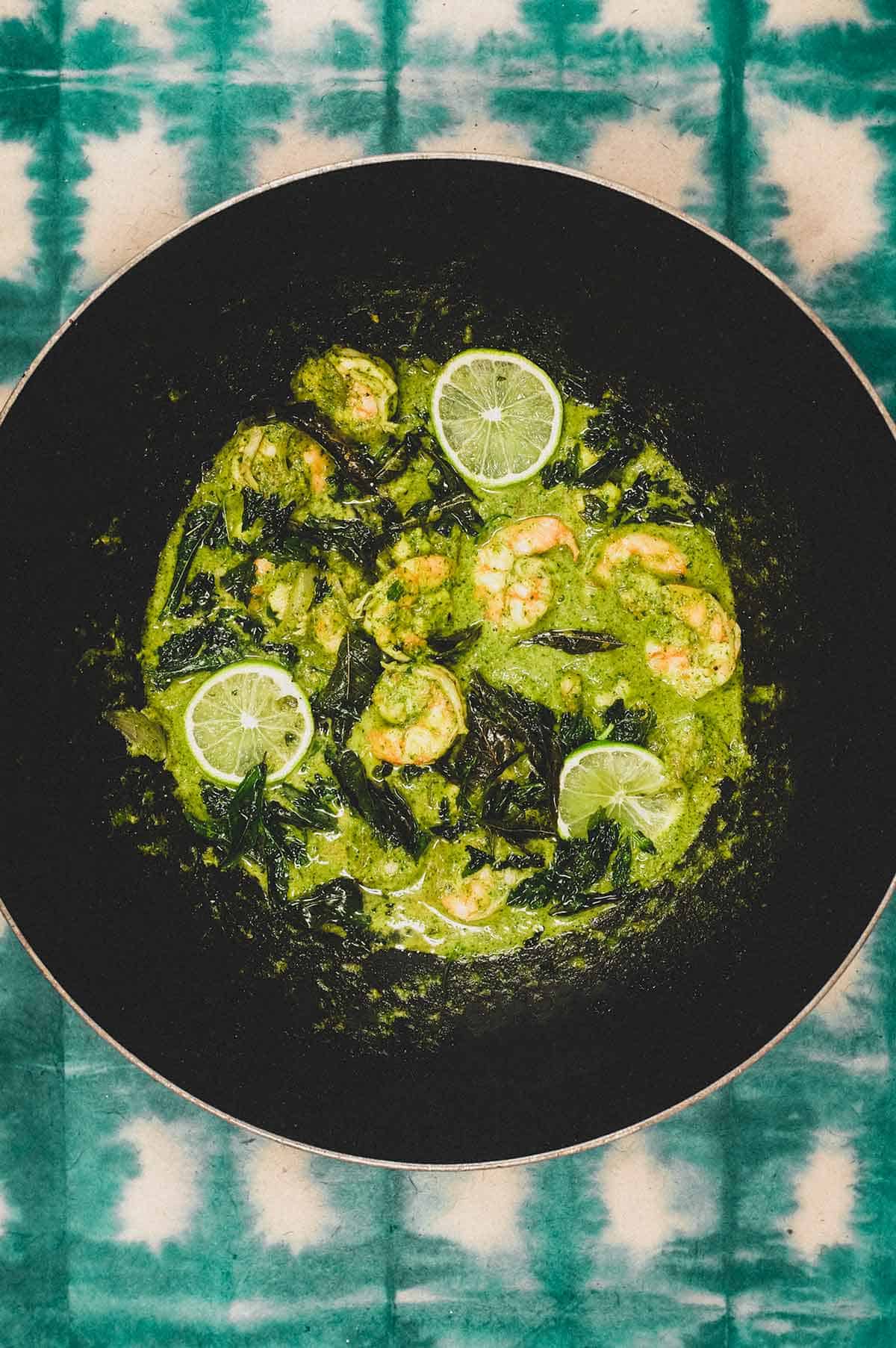 A pan of green masala with shrimp scattered with fried curry leaves and lime slices