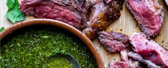 A small bowl of chimichurri sauce with a spoon sits alongside slices of rare cooked ribeye steak