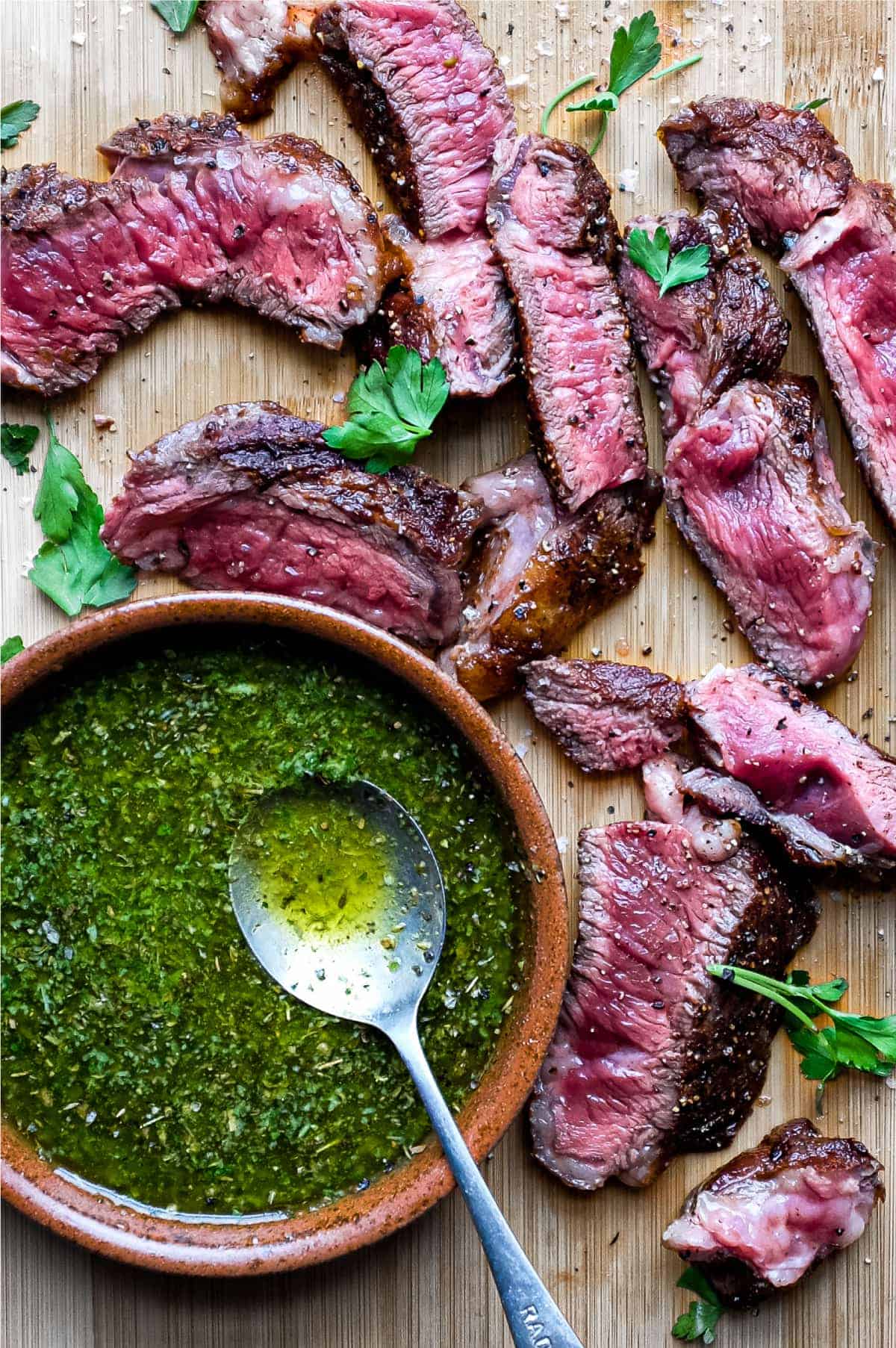 A small bowl of chimichurri sauce with a spoon sits alongside slices of rare cooked ribeye steak