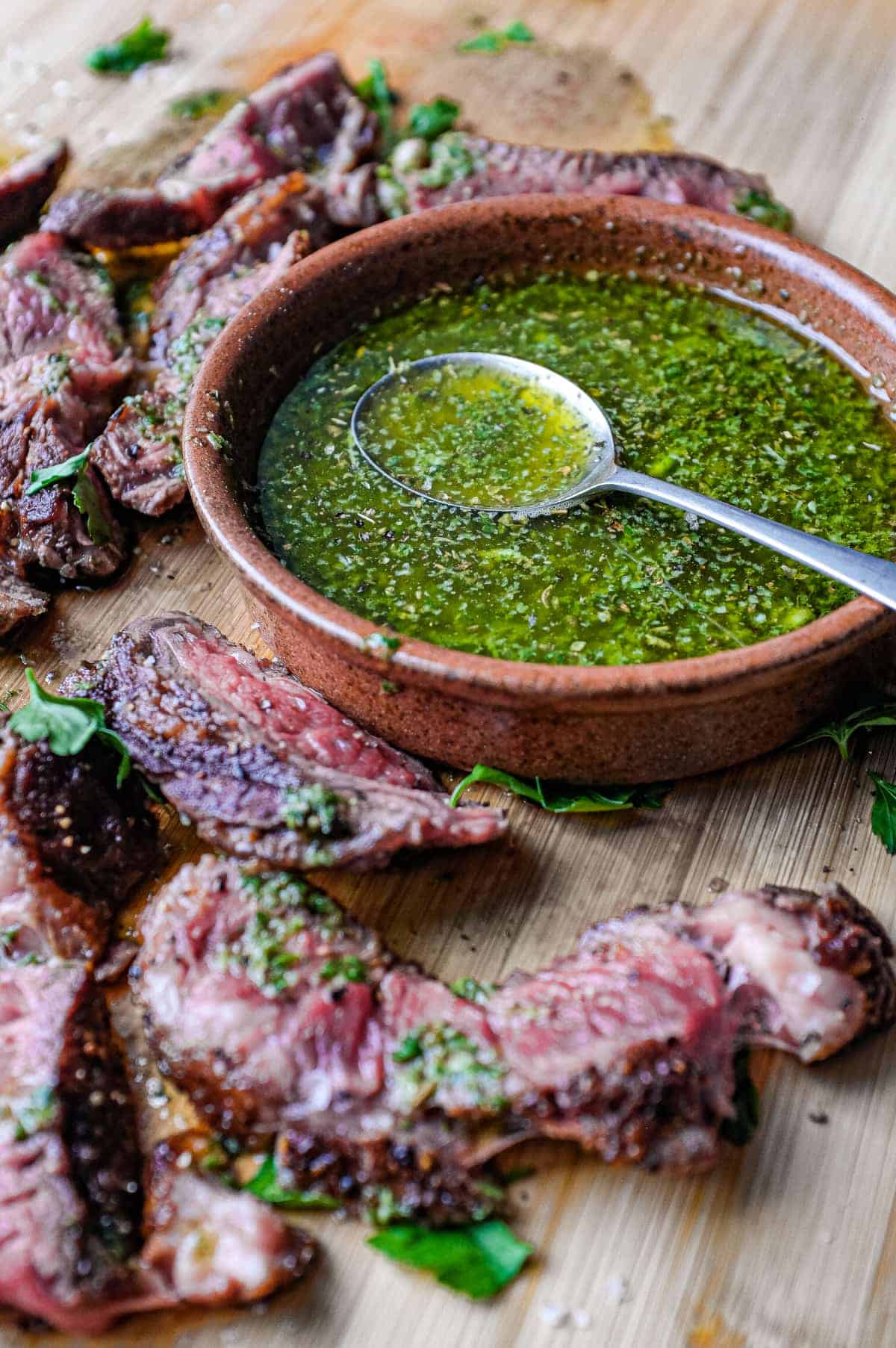 A small bowl of chimichurri sauce with a spoon sits alongside slices of rare cooked ribeye steak