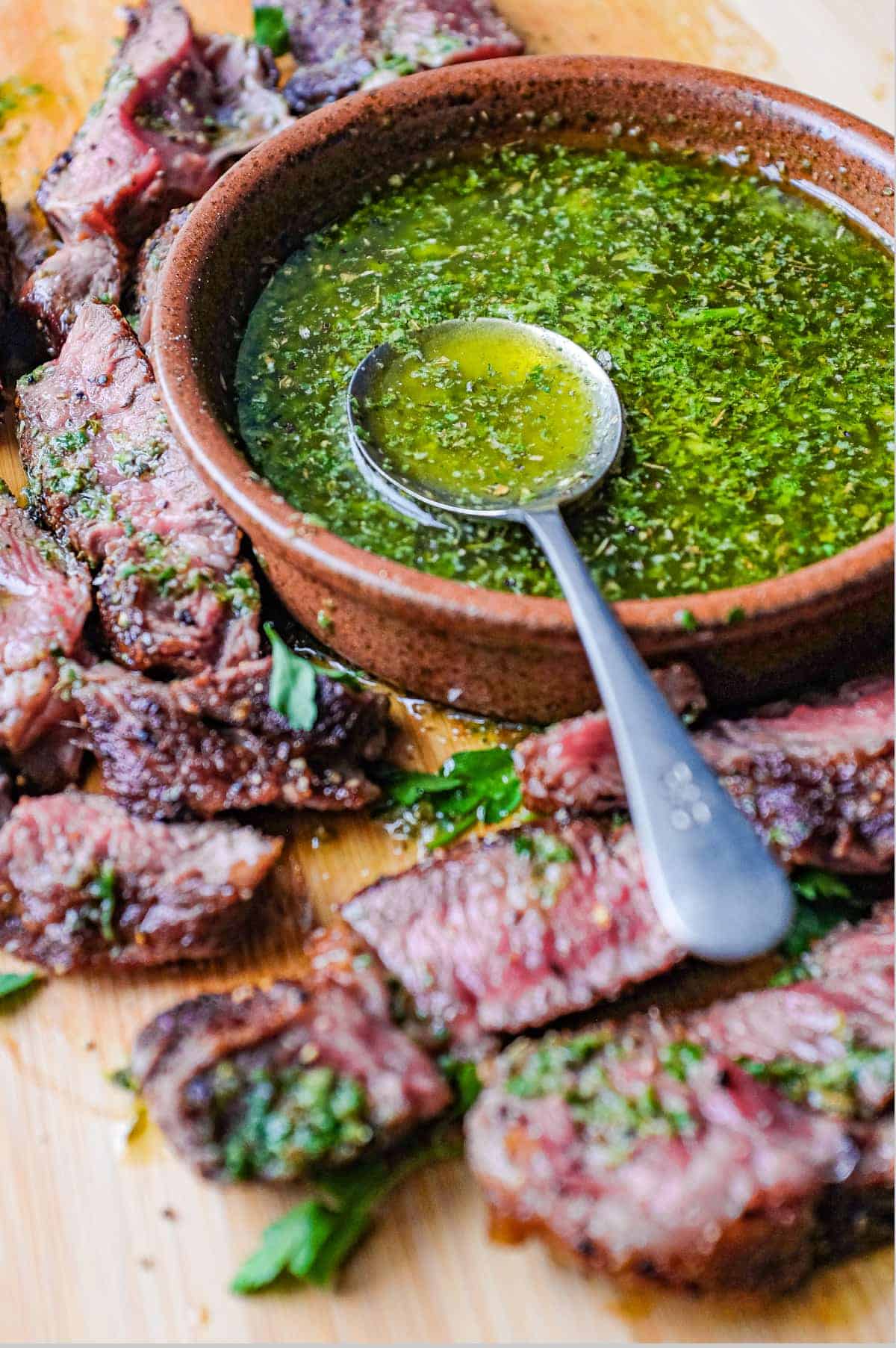 A small bowl of chimichurri sauce with a spoon sits alongside slices of rare cooked ribeye steak