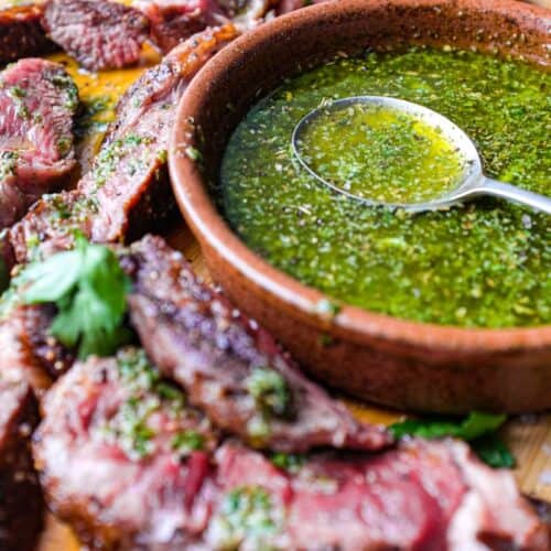A small bowl of chimichurri sauce with a spoon sits alongside slices of rare cooked ribeye steak