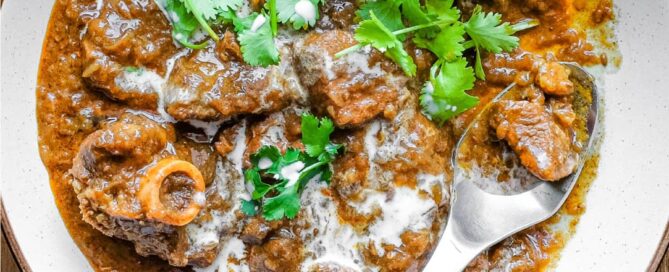 A bowl of lamb madras curry with a spoon on a wooden surface. The curry is garnished with cilantro and drizzled with coconut milk.
