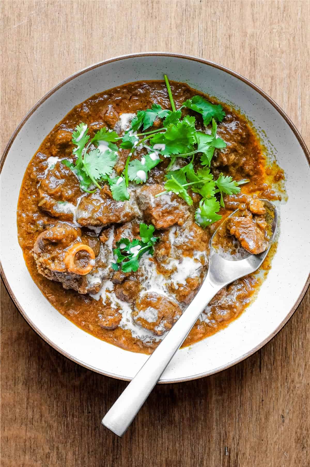 A bowl of lamb madras curry with a spoon on a wooden surface. The curry is garnished with cilantro and drizzled with coconut milk.