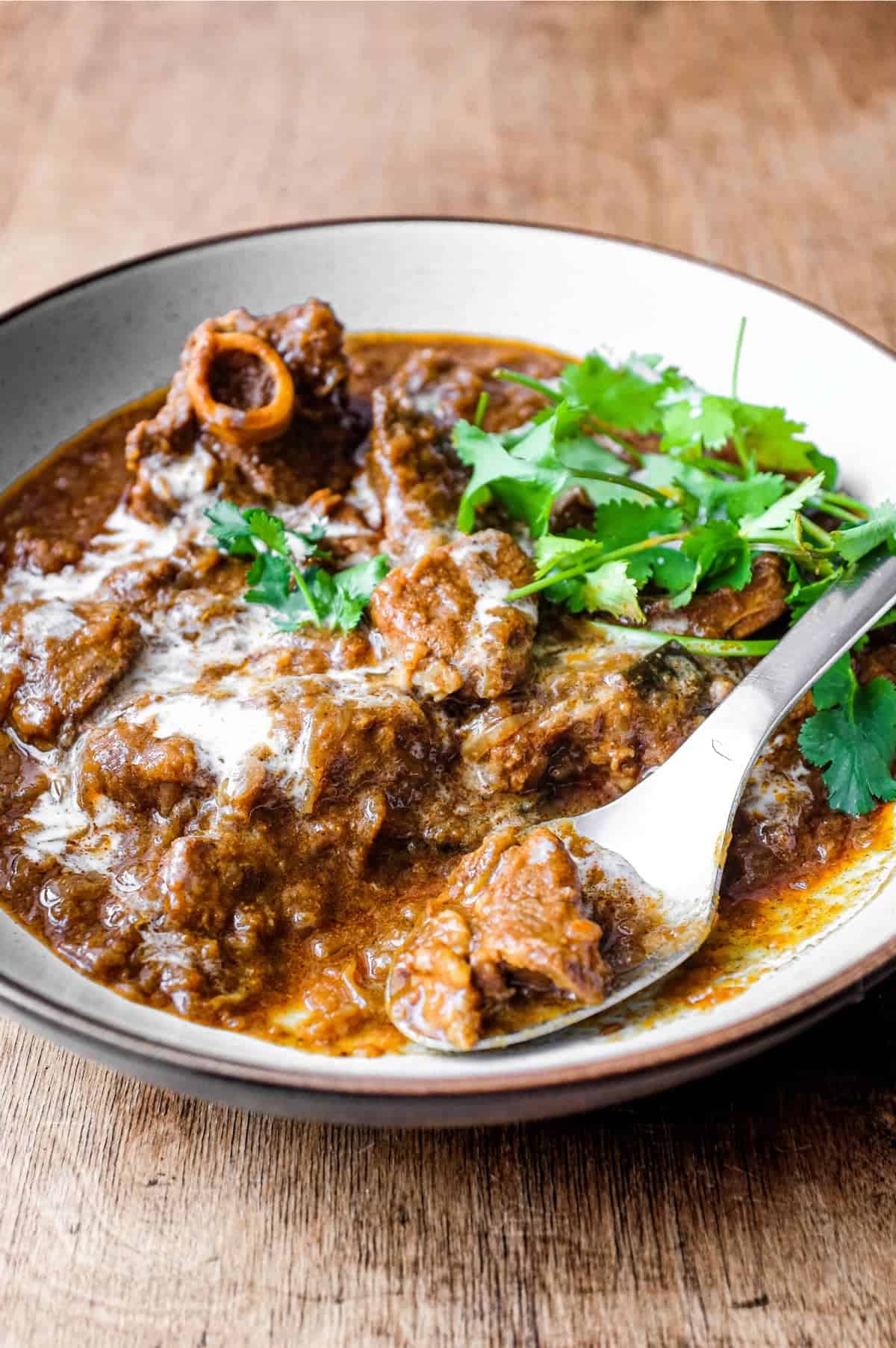 A bowl of lamb madras curry with a spoon on a wooden surface. The curry is garnished with cilantro and drizzled with coconut milk.