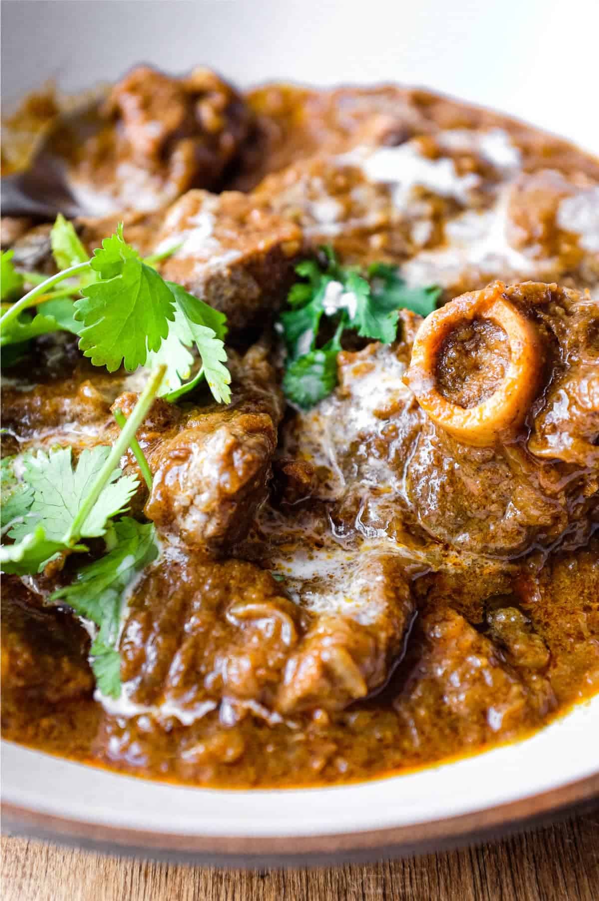 A bowl of lamb madras curry with a spoon on a wooden surface. The curry is garnished with cilantro and drizzled with coconut milk.