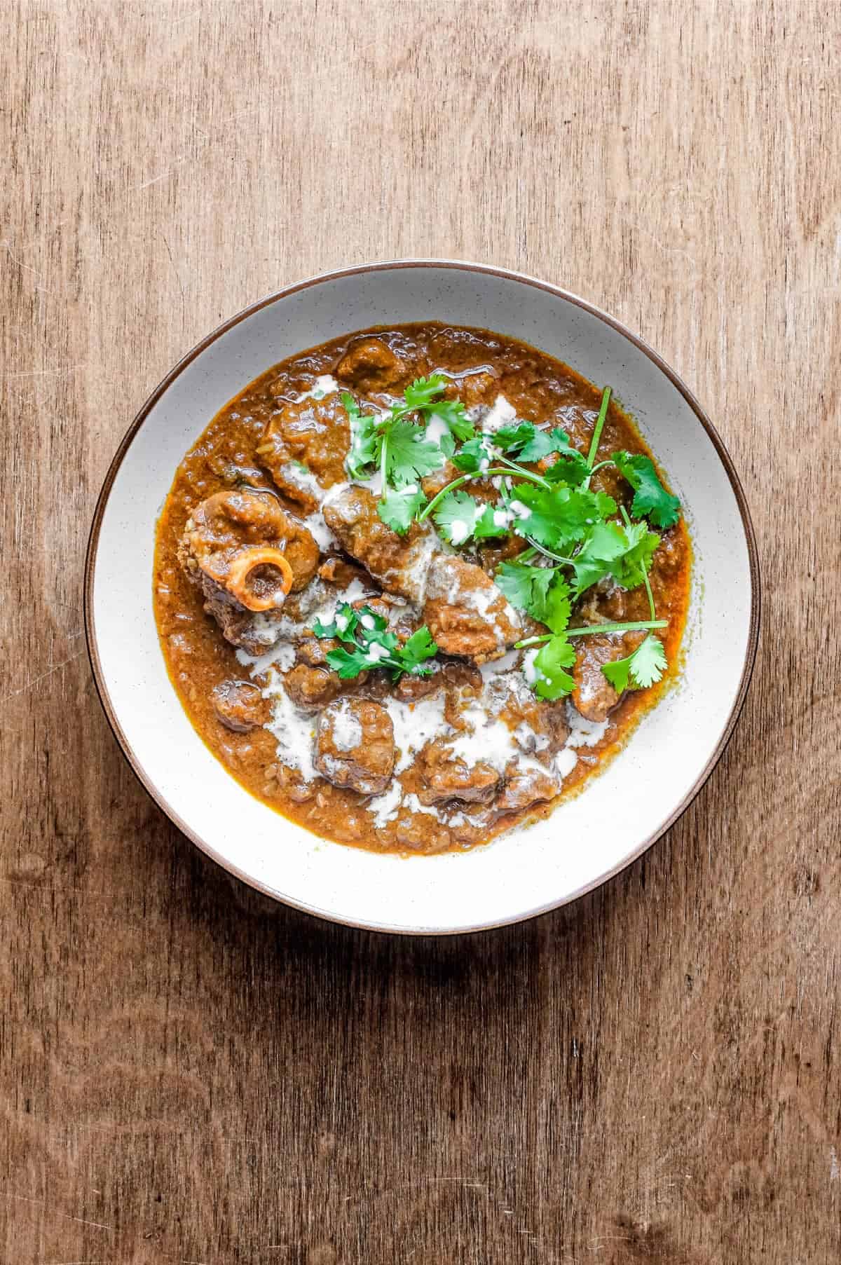 A bowl of lamb madras curry on a wooden surface. The curry is garnished with cilantro and drizzled with coconut milk.