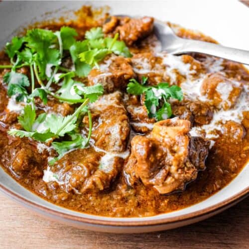 A bowl of lamb madras curry with a spoon on a wooden surface. The curry is garnished with cilantro and drizzled with coconut milk.