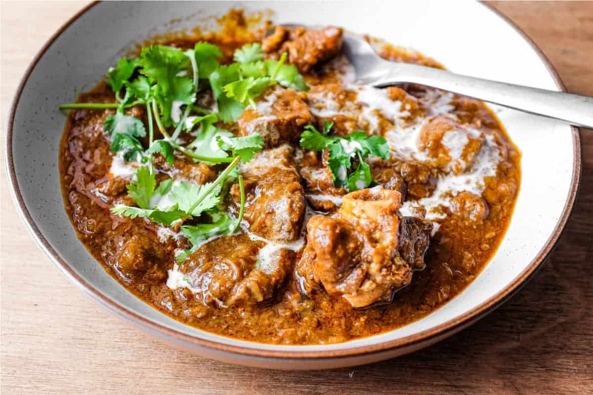 A bowl of lamb madras curry with a spoon on a wooden surface. The curry is garnished with cilantro and drizzled with coconut milk.