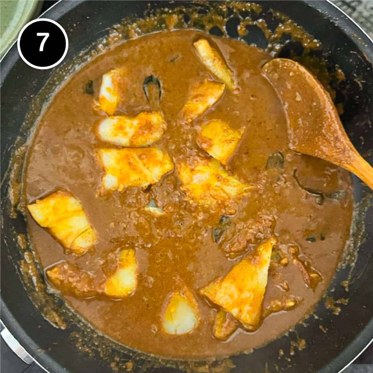 A pan of fish curry with a wooden spoon from above.