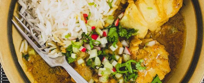 Bengali Fish Curry in a bowl in a black tabletop with rice and scattered with cilantro, radishes and celery