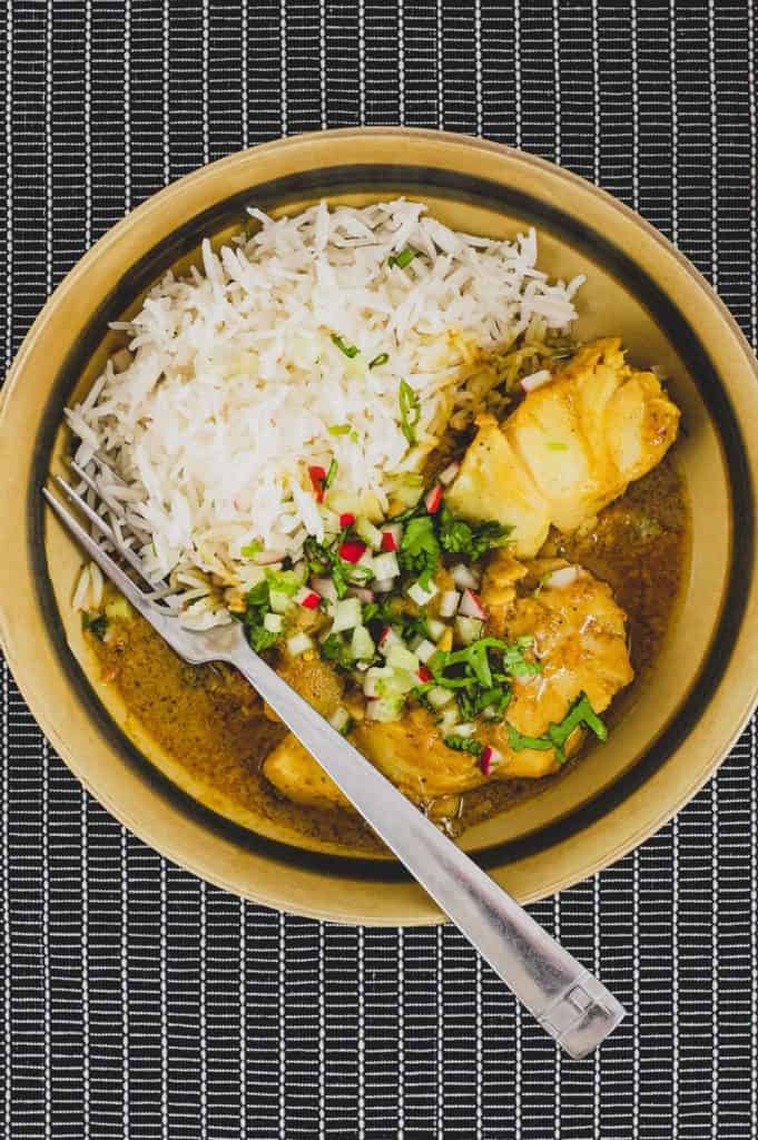 Bengali Fish Curry in a bowl in a black tabletop with rice and scattered with cilantro, radishes and celery