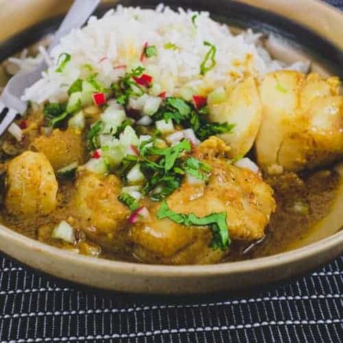 Bengali Fish Curry in a bowl in a black tabletop with rice and scattered with cilantro, radishes and celery