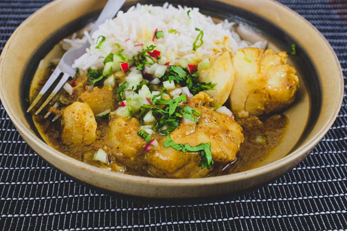 Bengali Fish Curry in a bowl in a black tabletop with rice and scattered with cilantro, radishes and celery