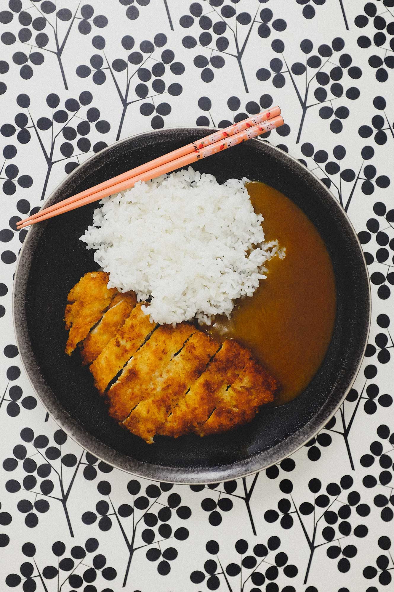 A plate of katsu curry with a katsu cutlet, rice and some Japanese-style curry sauce. Chopsticks sit on the side of the plate. There is a graphic black and white polka dot pattern in the background.