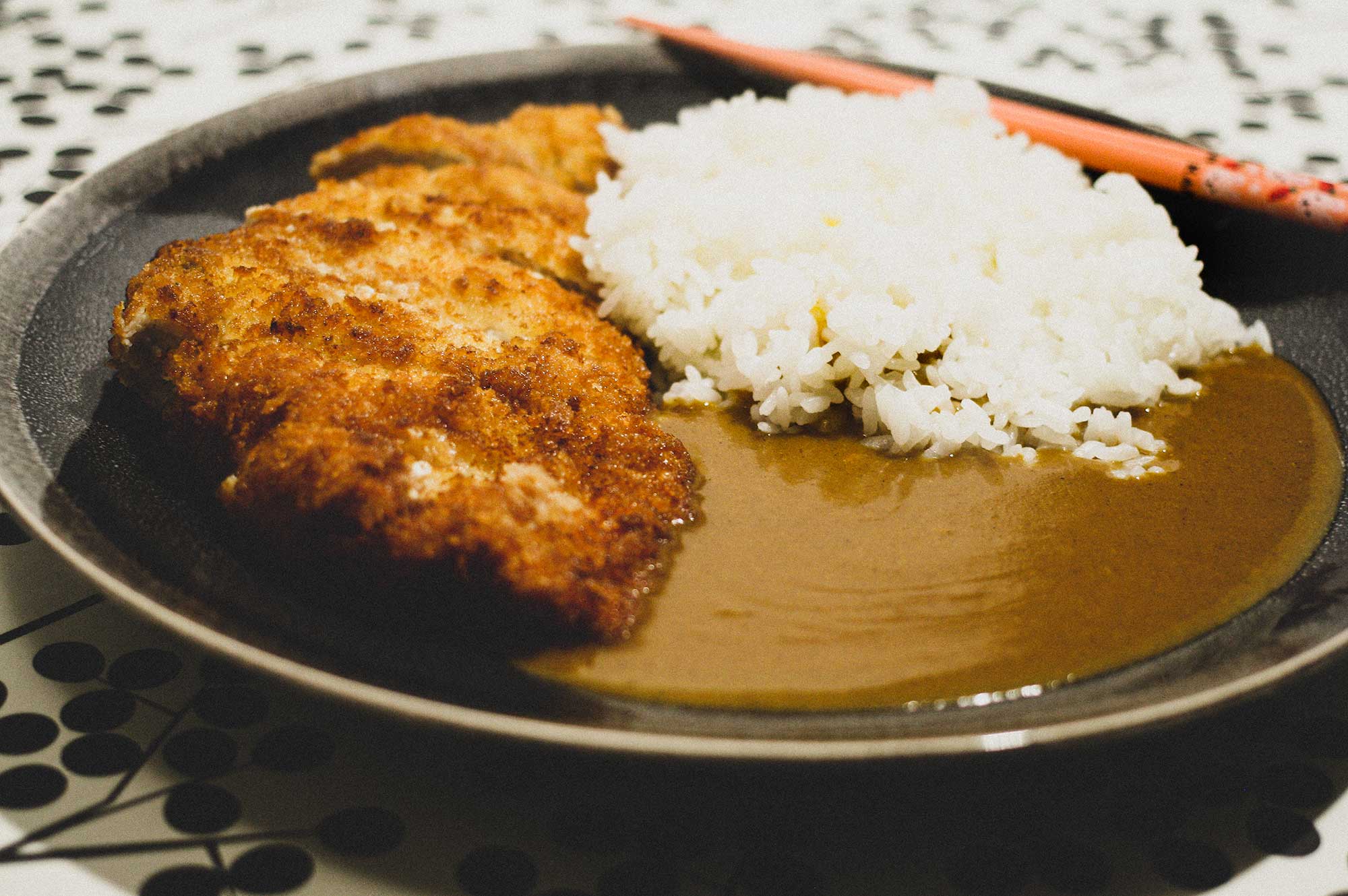 A plate of katsu curry with a katsu cutlet, rice and some Japanese-style curry sauce. Chopsticks sit on the side of the plate. There is a graphic black and white polka dot pattern in the background.