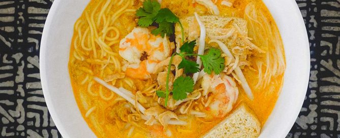 A bowl of Malaysian curry laksa decorated with prawns, tofu , beansprouts and herbs. Various condiments in small bowls sit by the side of the bowl along with chopsticks.