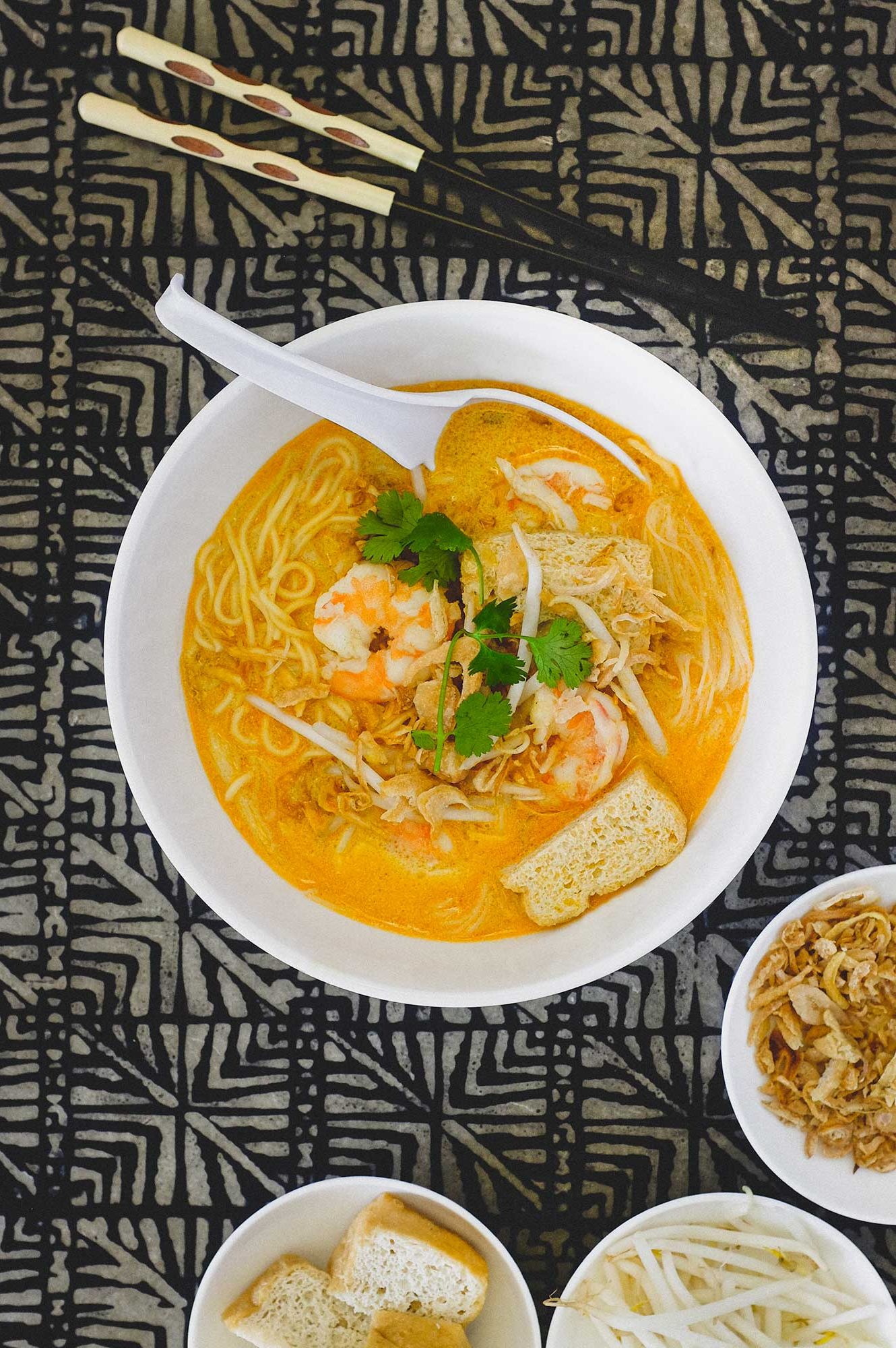 A bowl of Malaysian curry laksa decorated with prawns, tofu , beansprouts and herbs. Various condiments in small bowls sit by the side of the bowl along with chopsticks.