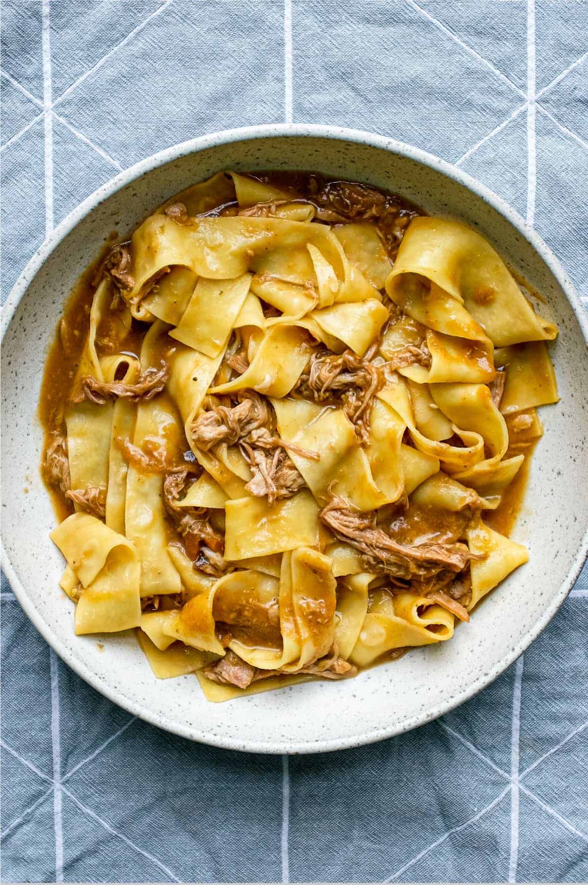 A bowl of pappardelle pasta with a lamb ragu sauce on a blue tea towel background