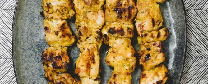 An oval platter holding four metal skewers of bbq chicken pieces. Known in Spain as Pinchos Morunos.