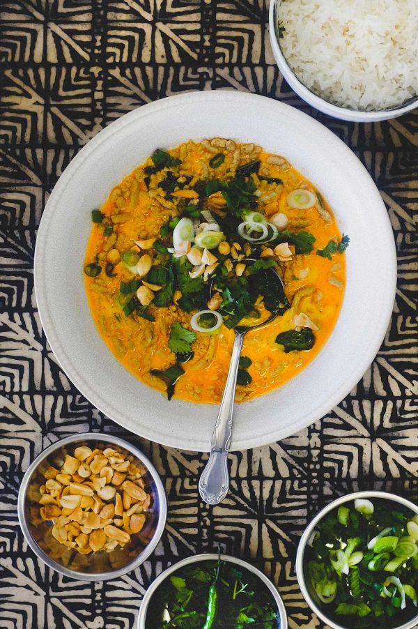 A bowl of Thai yellow curry with minced pork. Surrounding the bowl are smaller bowls of spring onions, cilantro, peanuts and jasmine rice.