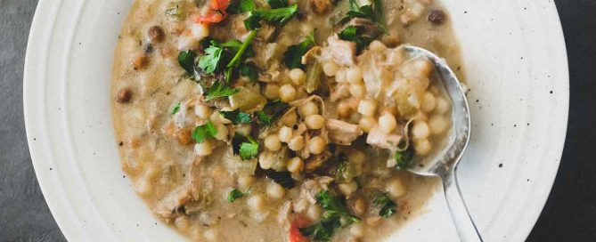 A bowl of chicken soup with fregola pasta and a spoon. Garnished with fresh herbs.