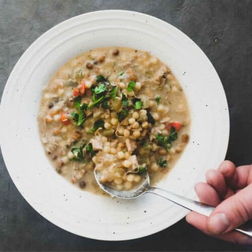A bowl of chicken soup with fregola pasta and a spoon. Garnished with fresh herbs.