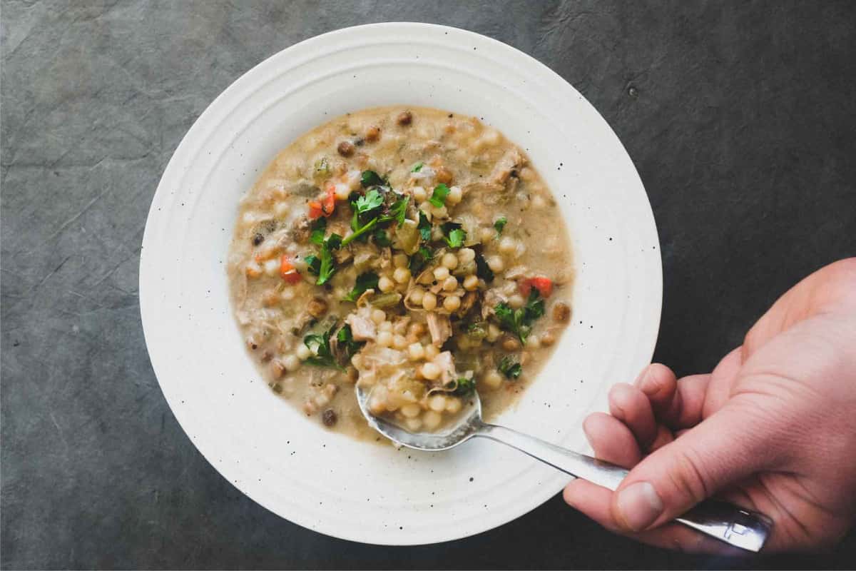 A bowl of chicken soup with fregola pasta and a spoon. Garnished with fresh herbs.