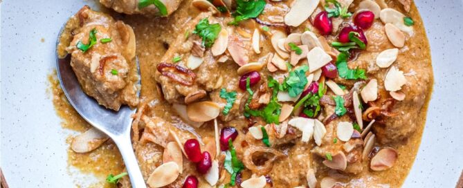 A bowl of Mughlai Chicken Curry garnished with Almonds, cilantro and pomegranate seeds with a spoon on the side.