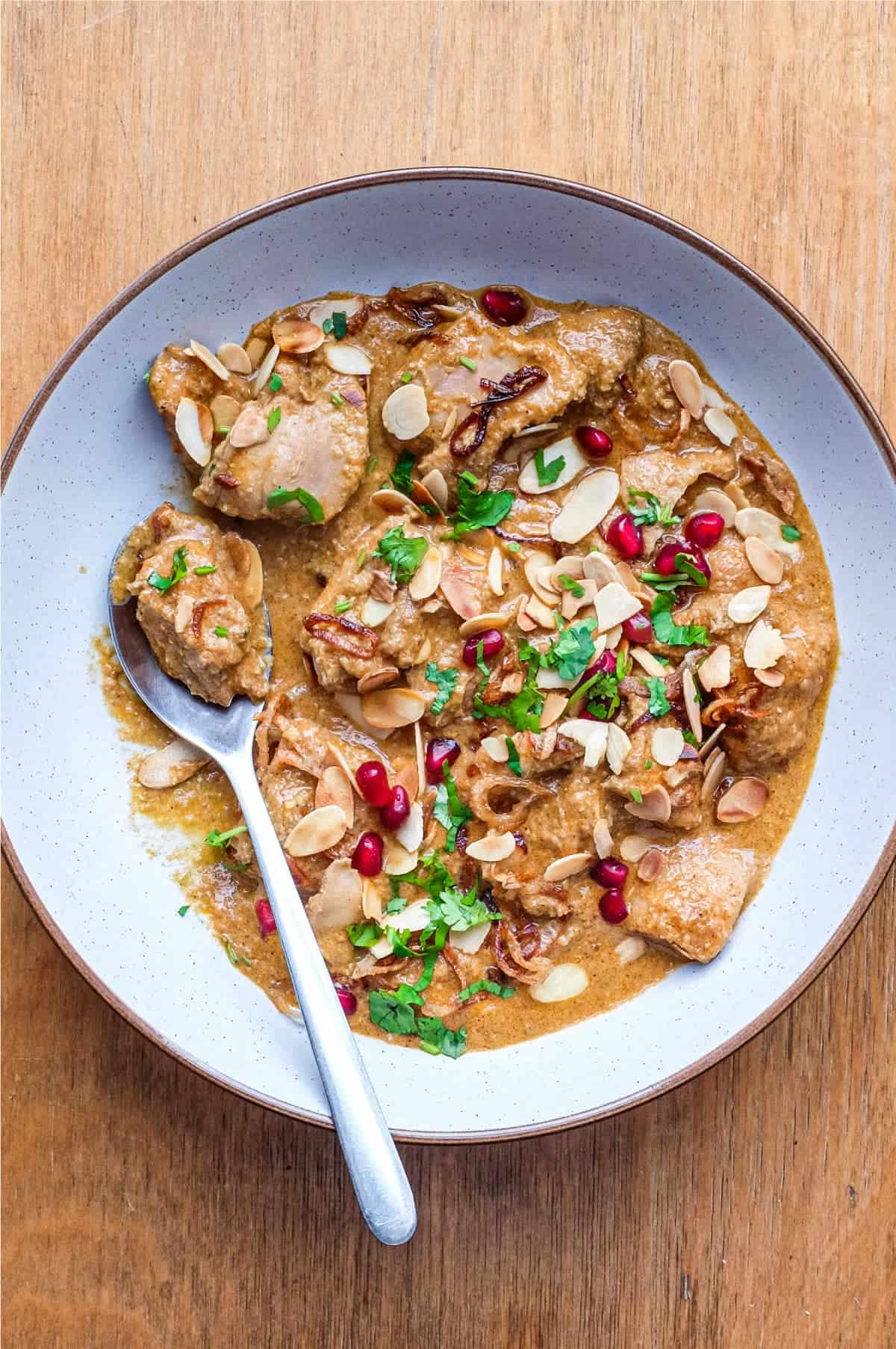 A bowl of Mughlai Chicken Curry garnished with Almonds, cilantro and pomegranate seeds with a spoon on the side.