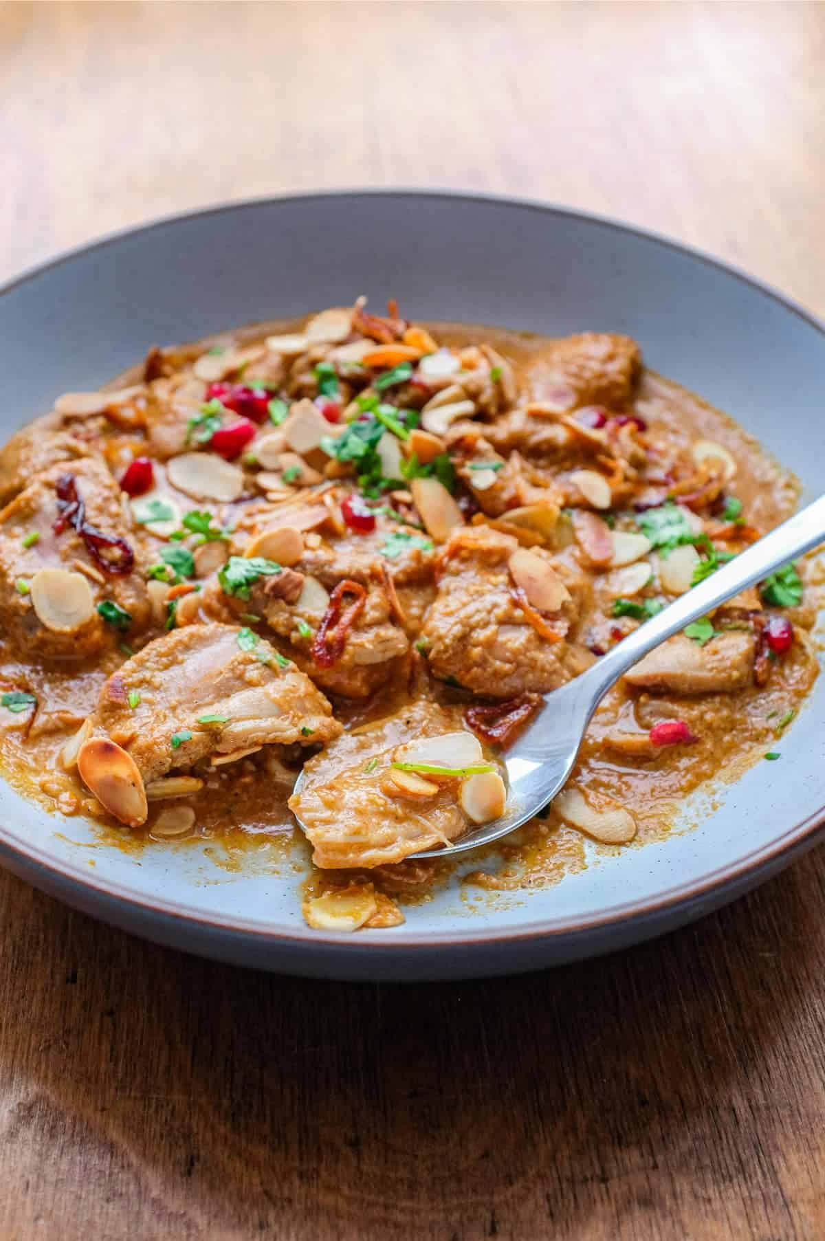 A bowl of Mughlai Chicken Curry garnished with Almonds, cilantro and pomegranate seeds with a spoon on the side.