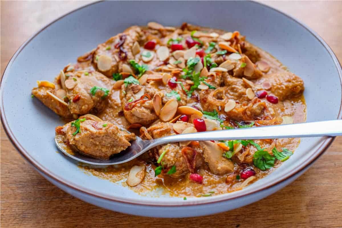 A bowl of Mughlai Chicken Curry garnished with Almonds, cilantro and pomegranate seeds with a spoon on the side.