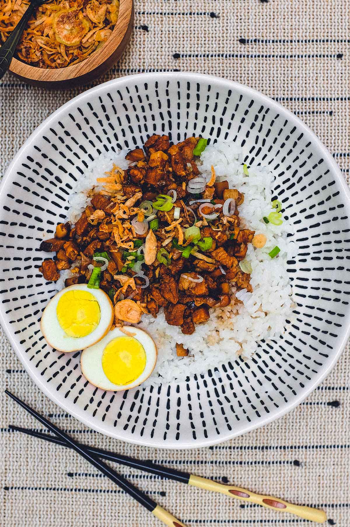 A bowl of Taiwanese braised pork (Lu Rou Fan) on a bed of rice with two egg halves, crispy onion and spring onions as a garnish.