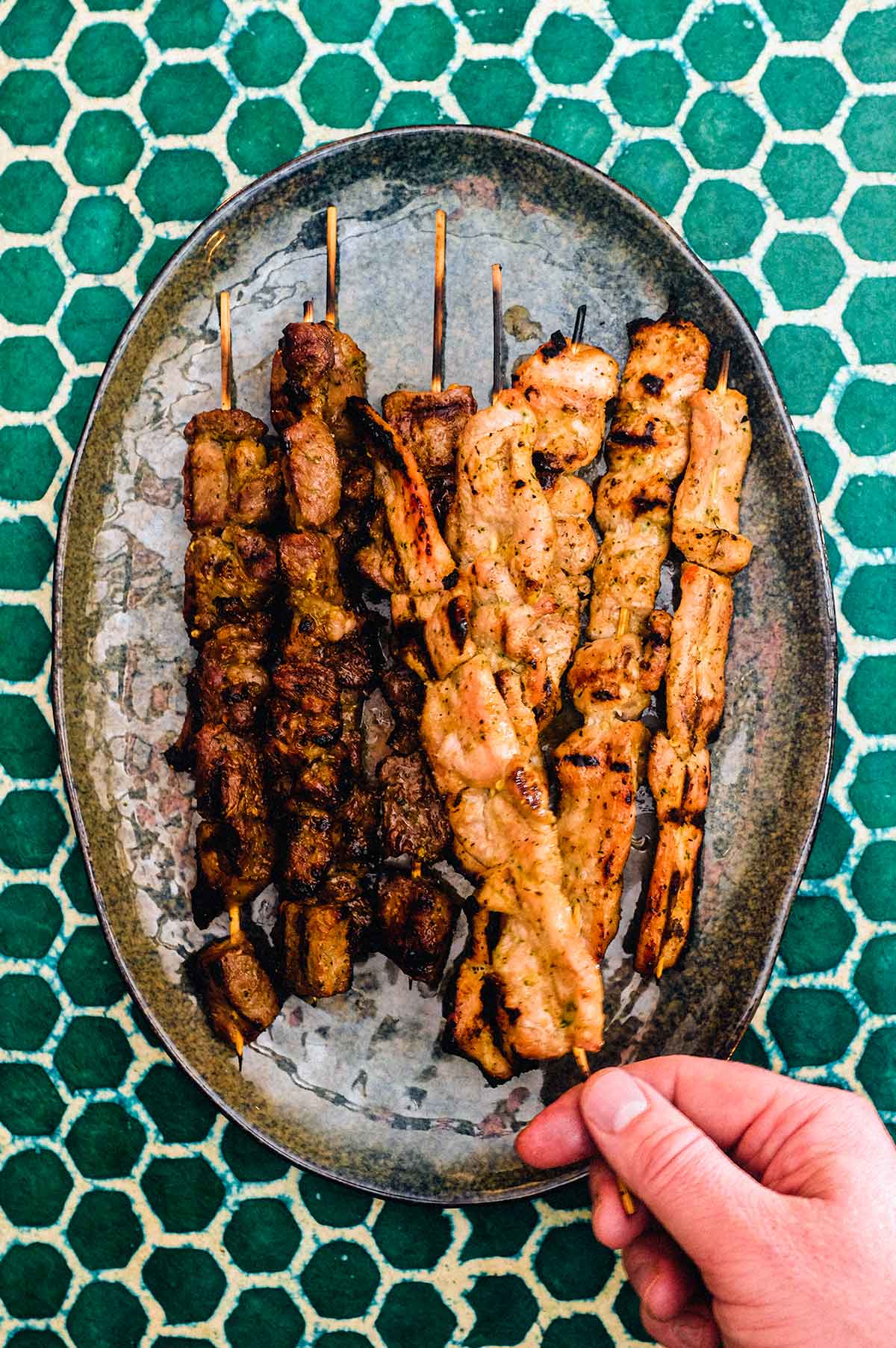 Beef and Pork satay skewers sit on a serving platter on a green batik background.