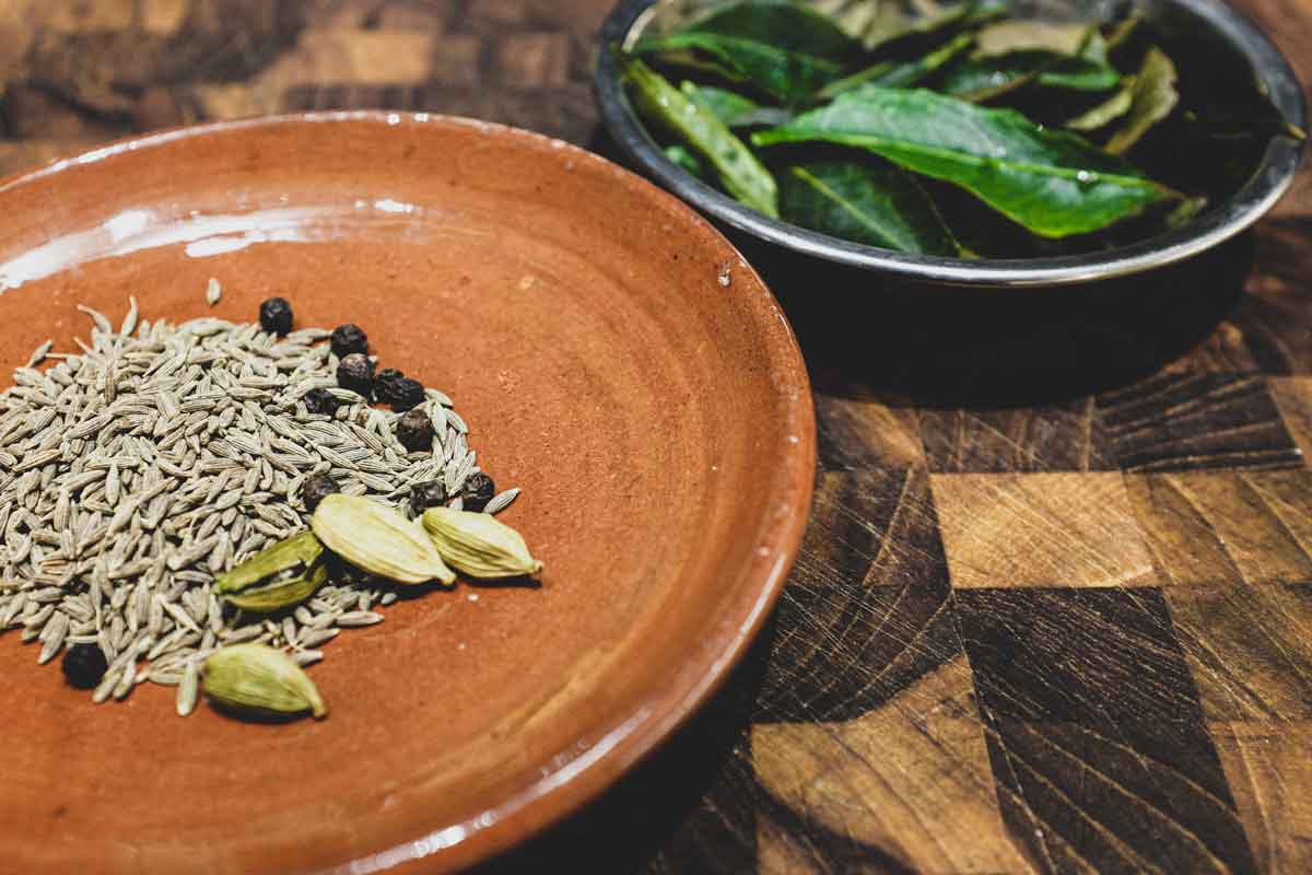 whole spices (cumin seeds, peppercorns and cardamom pods) on a small pottery plate with fresh curry leaves beside.