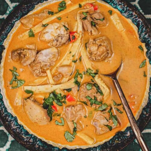 A bowl of vibrant orange Laotian chicken curry and spoon on a green batik background 