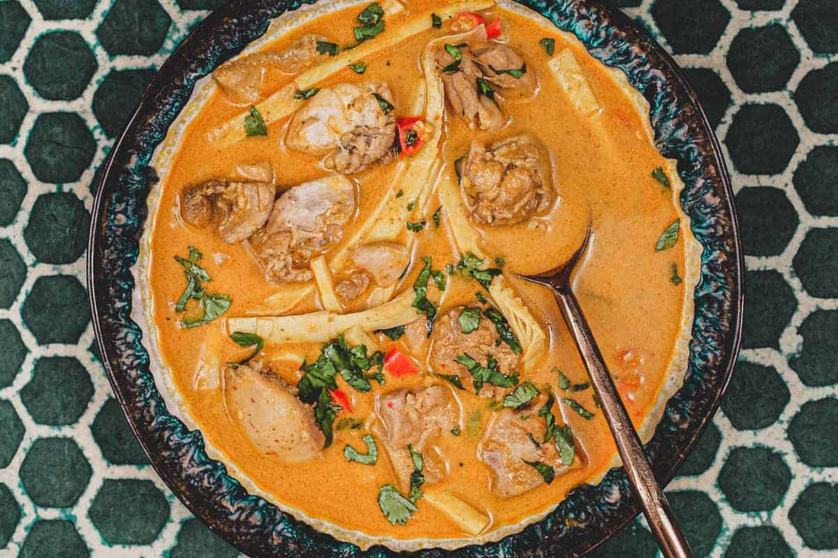 A bowl of vibrant orange Laotian chicken curry and spoon on a green batik background 