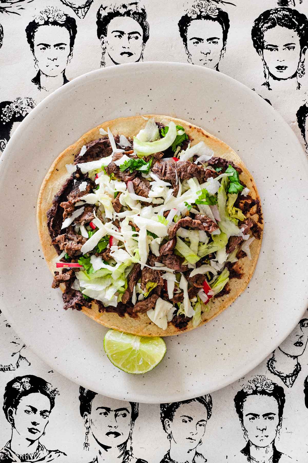 A plate with a Mexican tlayudas and wedge of lime. The background is a collage of sketches of Mexican artist Frida Kahlo