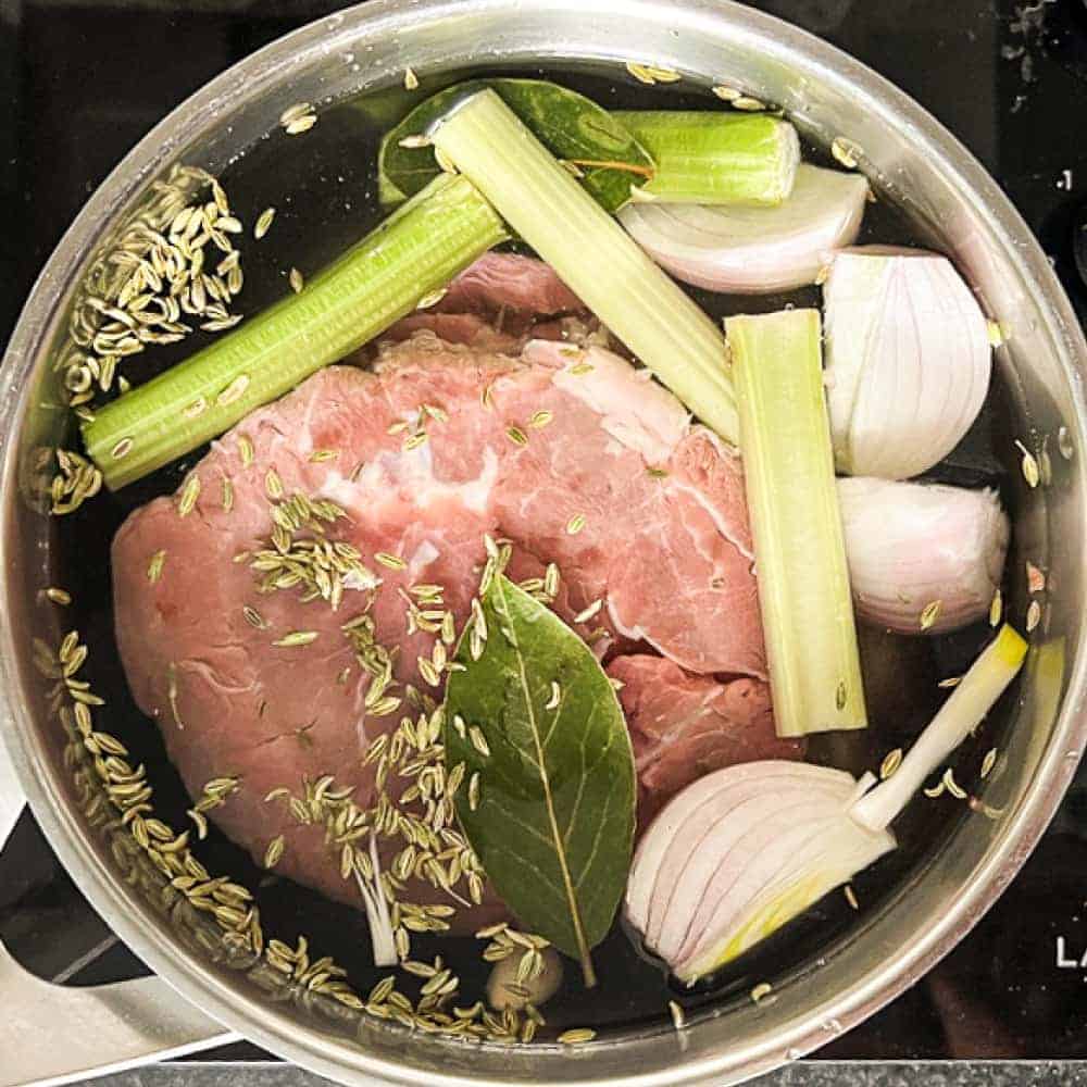 A shoulder of veal sits in a pan of water with celery, garlic, bay leaves, peppercorns and fennel seeds.