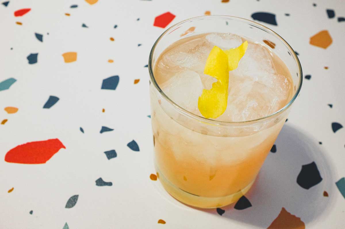 A highball glass with a Pamplemousse Cocktail on a stylish terrazzo surface. A twist of lemon rind sits on top.