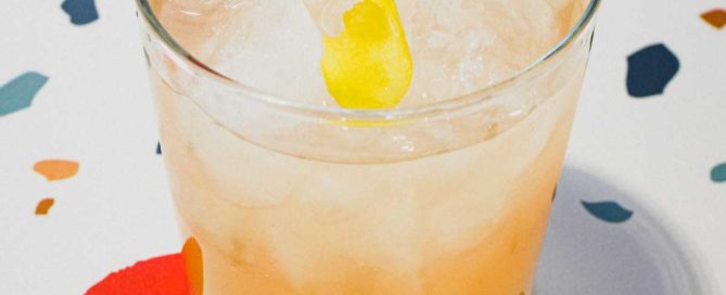 A highball glass with a Pamplemousse Cocktail on a stylish terrazzo surface. A twist of lemon rind sits on top.