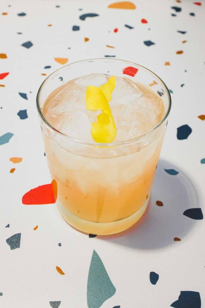 A highball glass with a Pamplemousse Cocktail on a stylish terrazzo surface. A twist of lemon rind sits on top.