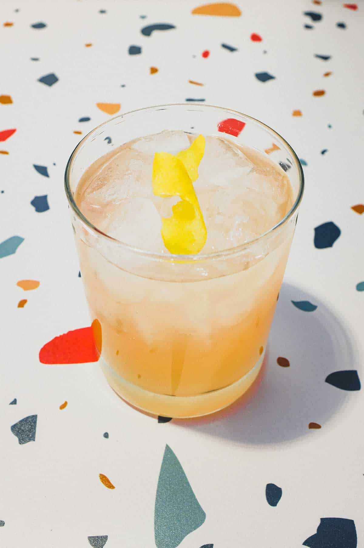 A highball glass with a Pamplemousse Cocktail on a stylish terrazzo surface. A twist of lemon rind sits on top.