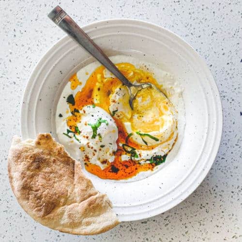 A small bowl of Turkish Cilbir (poached eggs) drizzled in orange Aleppo pepper butter with a piece of flatbread