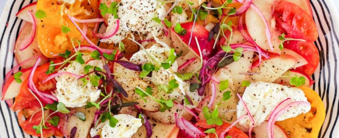 A salad of peaches, burrata cheese, tomato, red onion and micro greens sits on a marble countertop