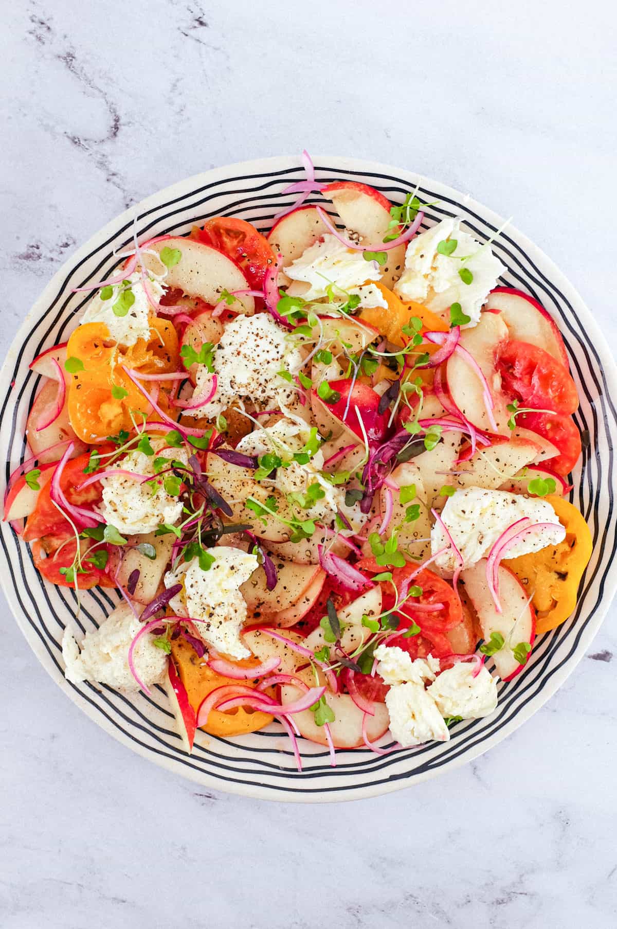 A salad of peaches, burrata cheese, tomato, red onion and micro greens sits on a marble countertop