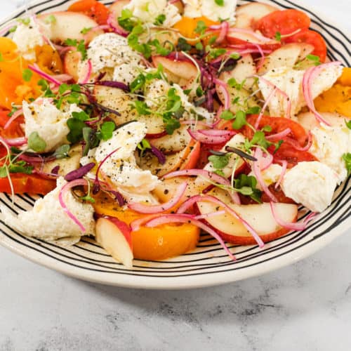 A salad of peaches, burrata cheese, tomato, red onion and micro greens sits on a marble countertop